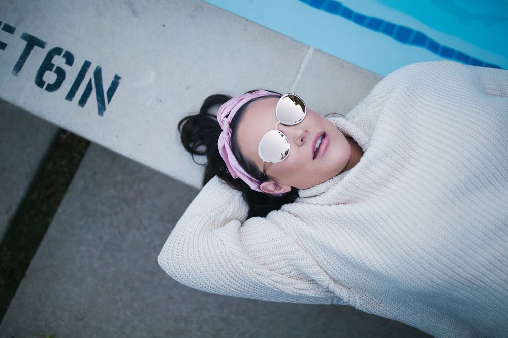 woman with mirrored sunglasses laying on edge of bright blue pool in sweater, vintage pink headband, with retro makeup