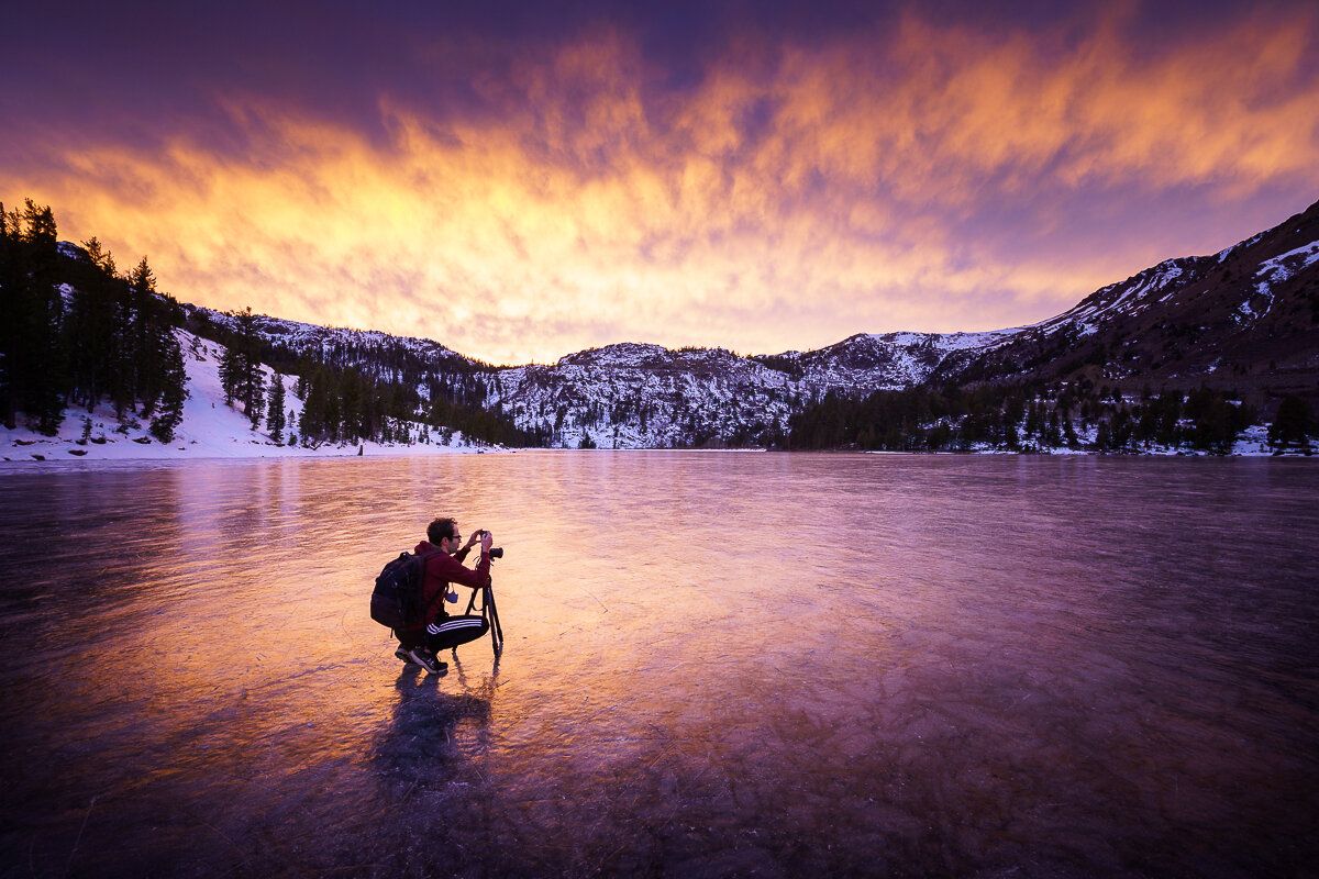 A photographer (Jeff) silhouetted against a frozen lake at sunset with snow-capped mountains and dramatic pink clouds in background.