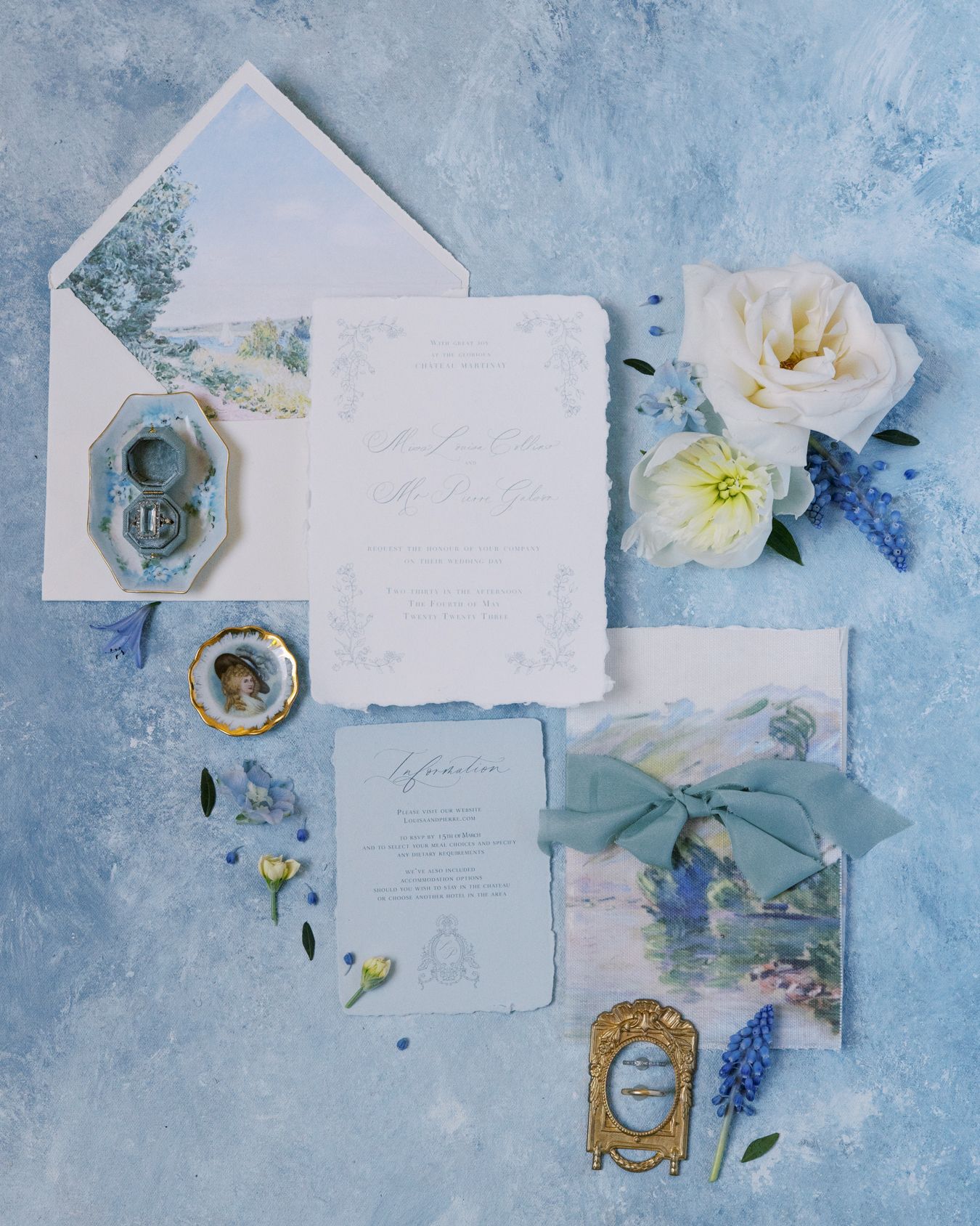 Wedding stationery flatlay with delicate white flowers, blue accents, and vintage frames on a textured blue background.