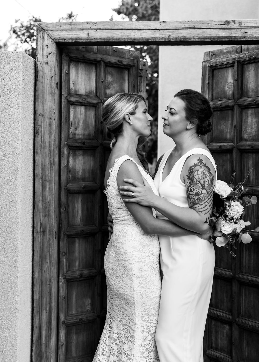 Two brides in white dresses share a romantic moment against rustic wooden doors.