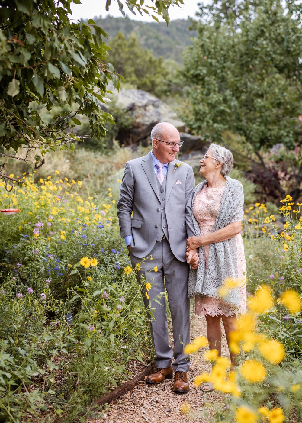 A couple stands together on a garden path surrounded by yellow wildflowers in a romantic outdoor setting.