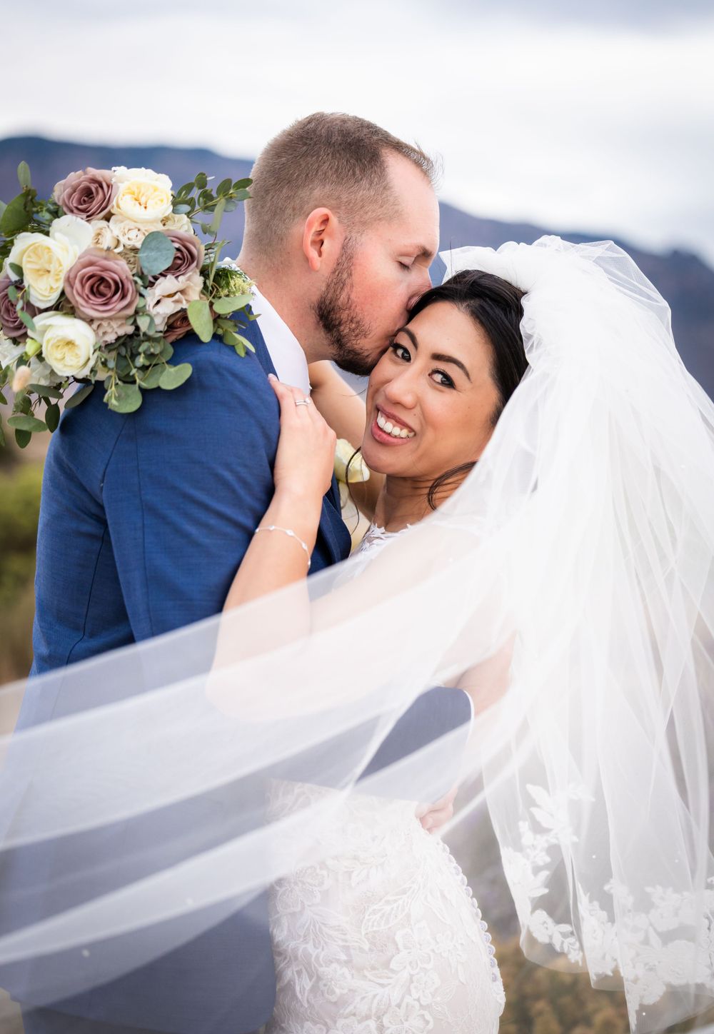 A tender wedding moment with a bride in white dress and veil being kissed while holding a floral bouquet.