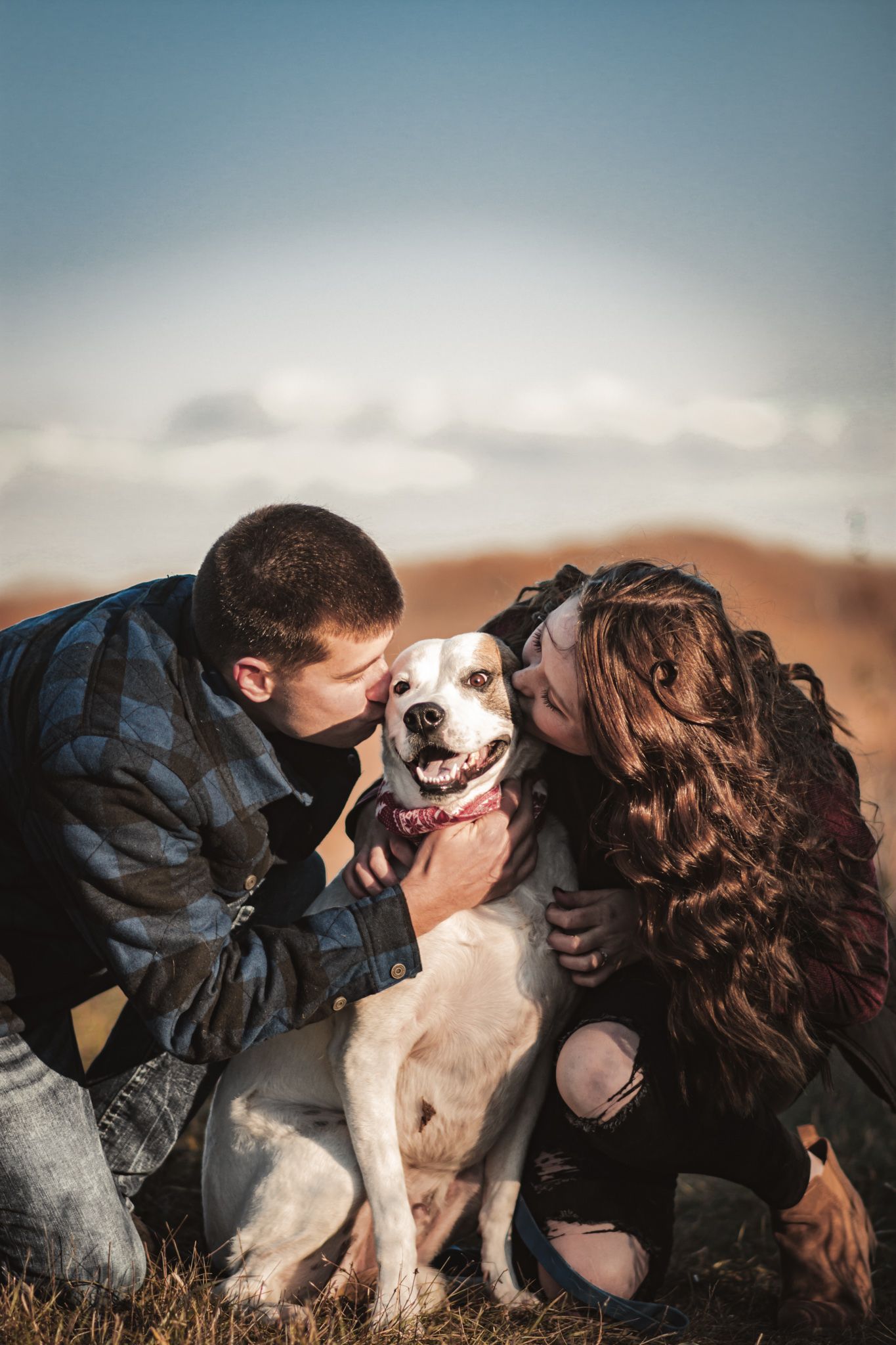 Couple embracing their smiling white dog in outdoor casual portrait.