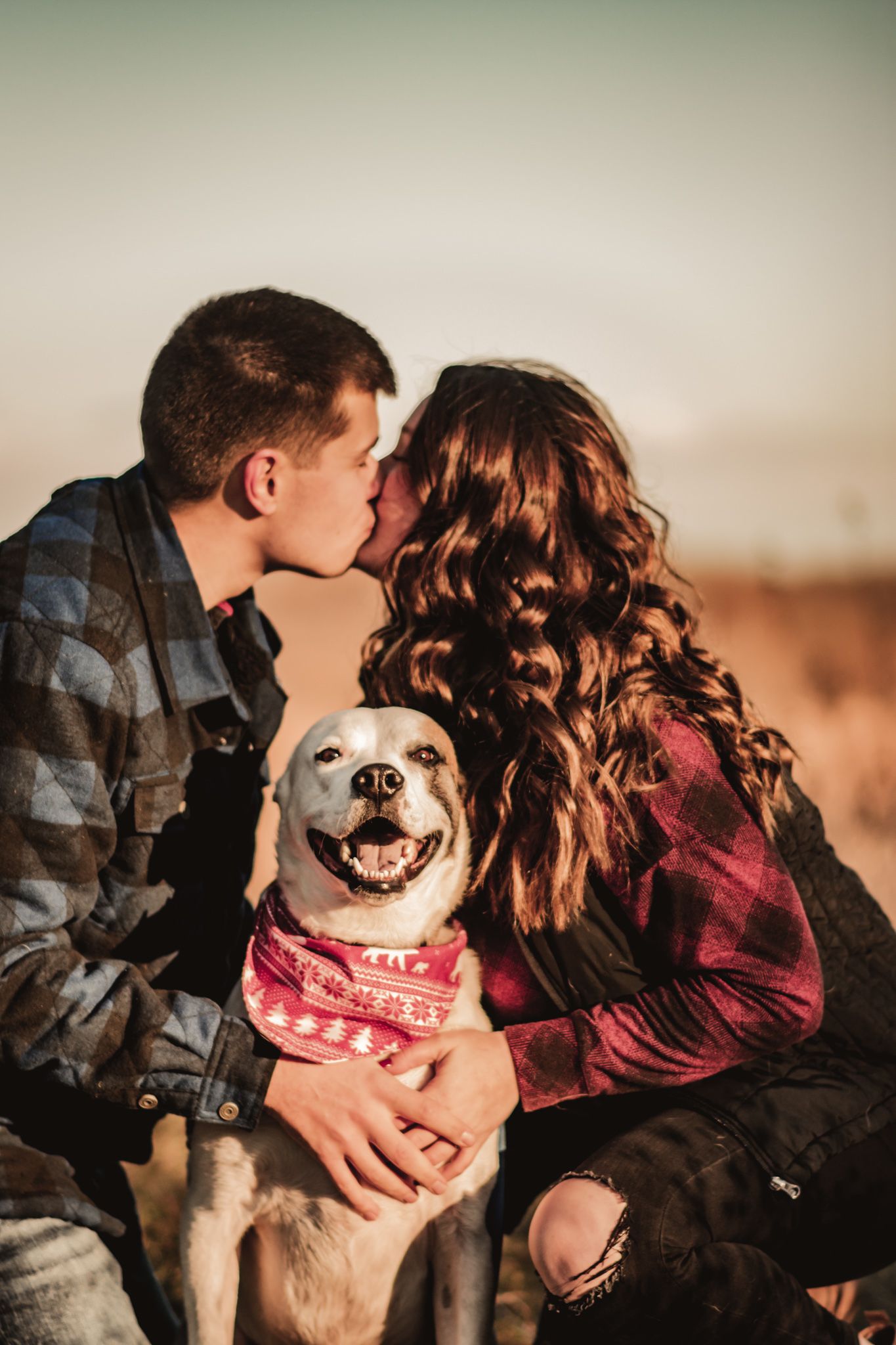 Couple sharing intimate moment with happy white dog wearing bandana at sunset.