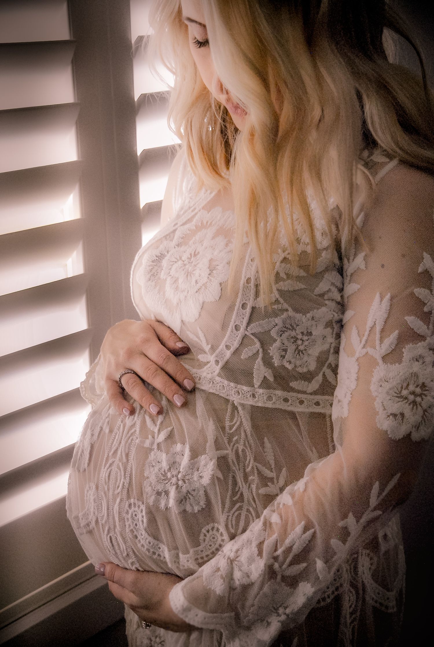 Delicate white lace maternity dress photographed by window light with soft shadows.