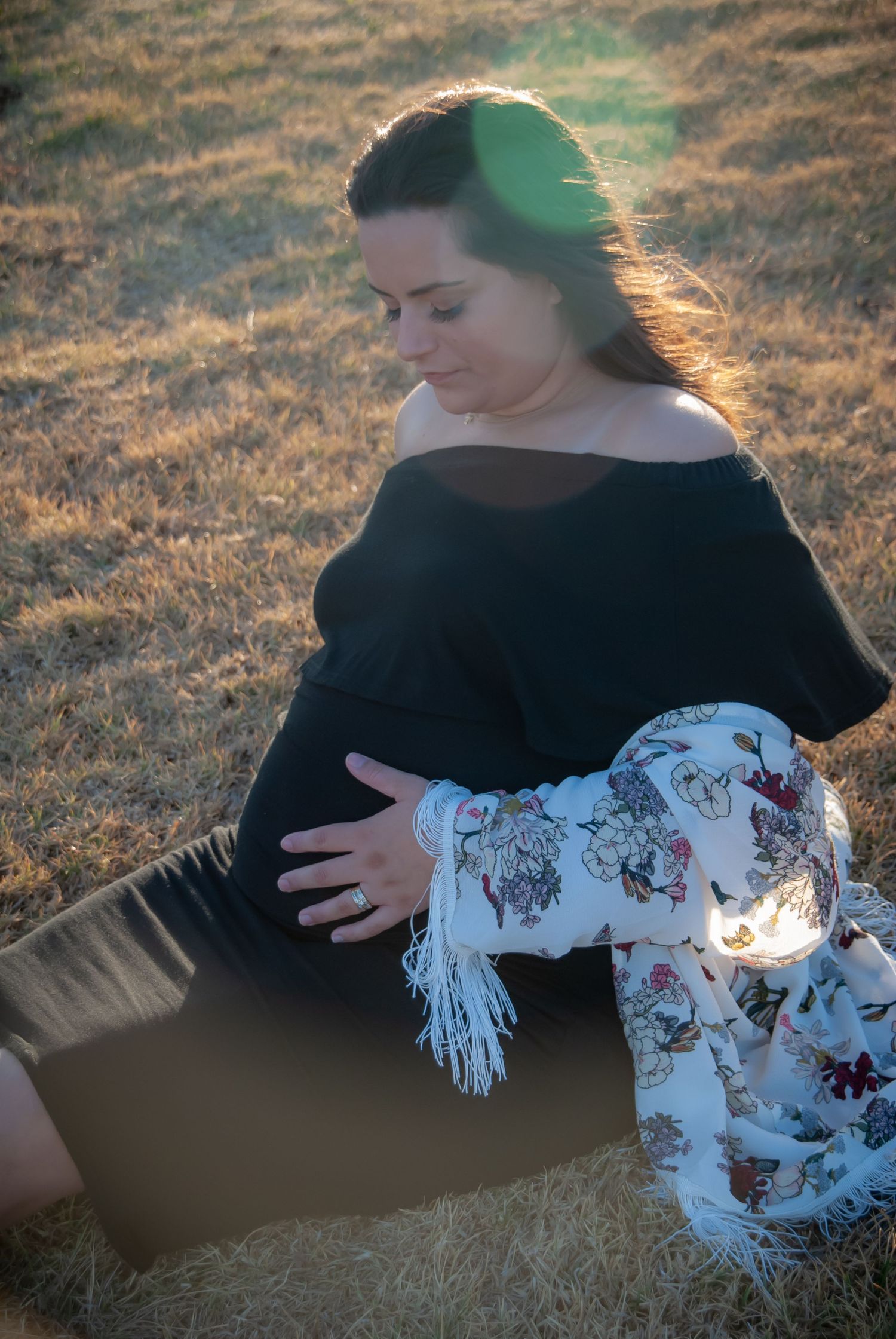 Woman in black dress sits outdoors at sunset holding a decorative floral pillow.