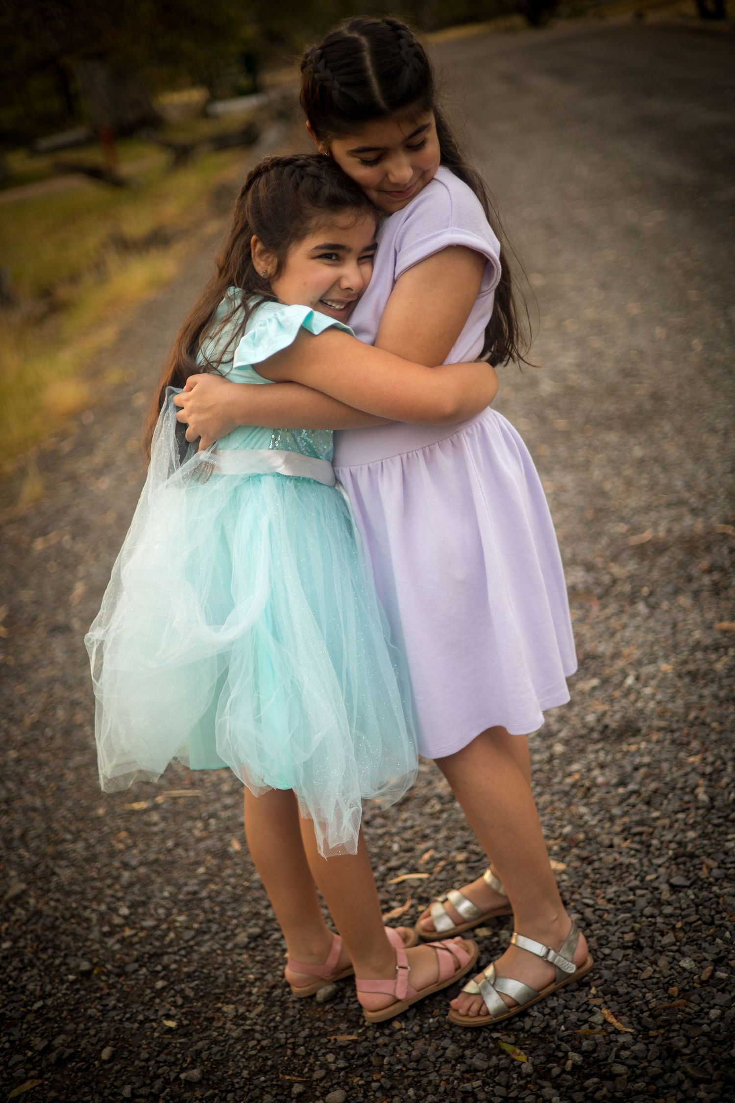 Two young figures in party dresses share a sweet embrace on a gravel path.