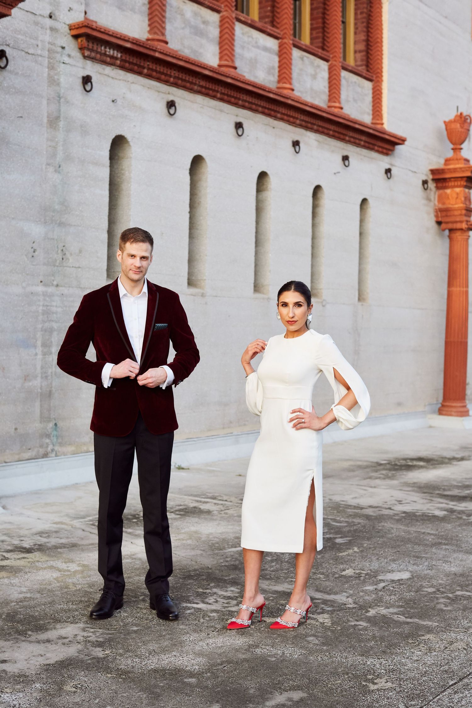 Stylish couple in formal attire posing against white building with architectural columns.