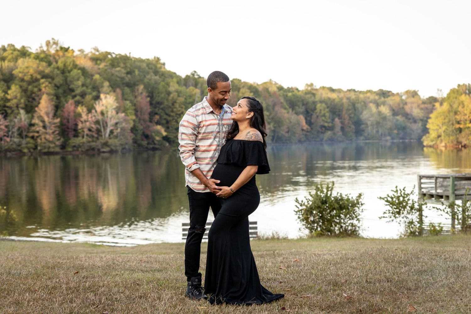 Maternity moment by serene lake with couple in black dress and striped shirt.