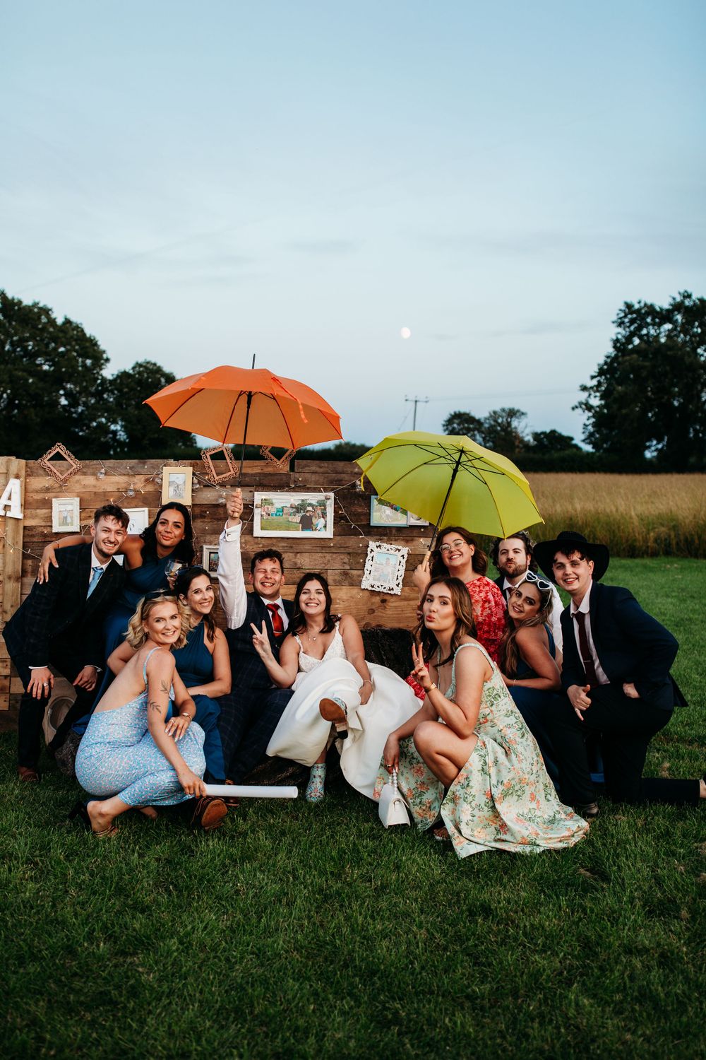 Wedding party poses with colorful umbrellas against rustic wooden backdrop in outdoor setting.