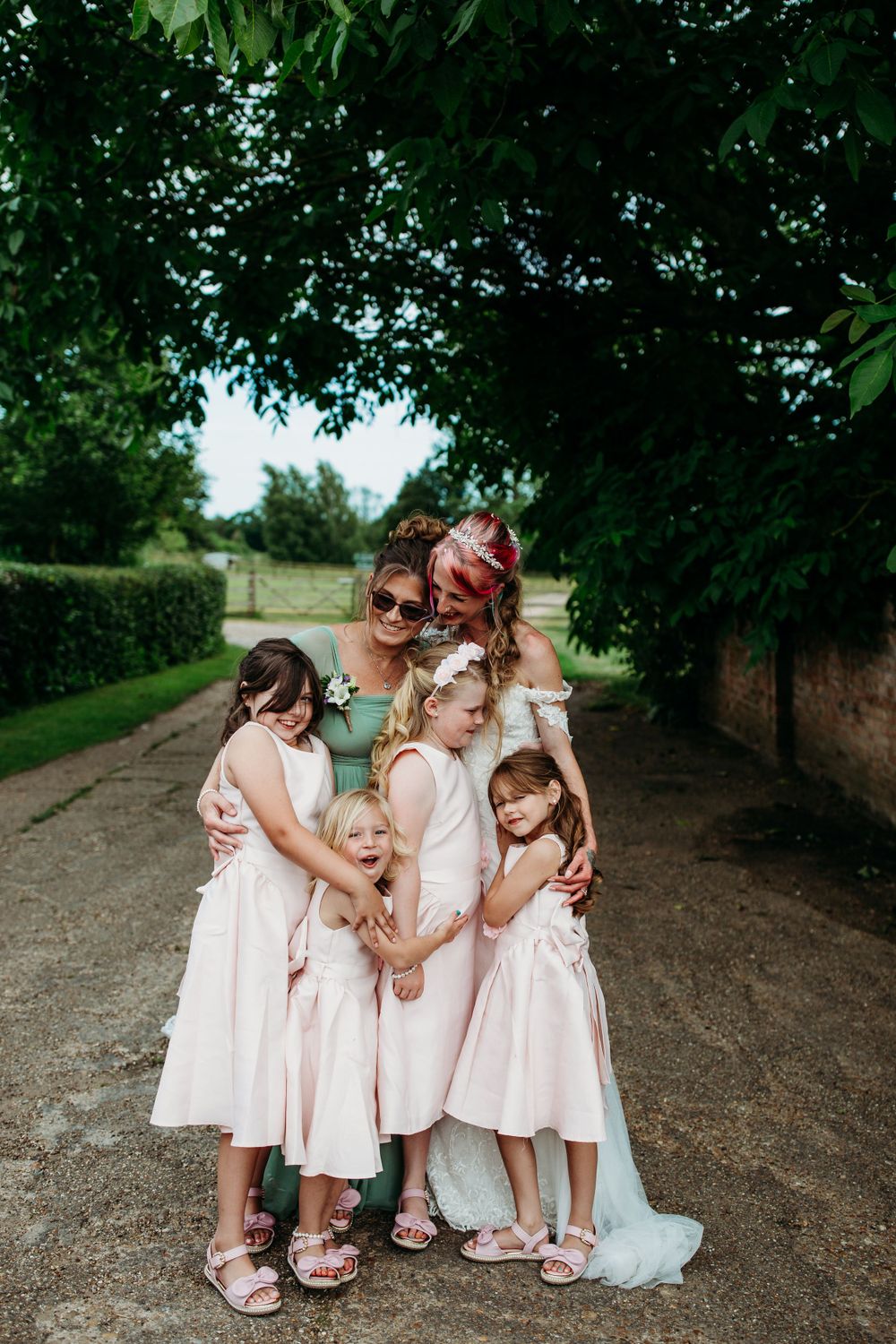 Group hug of young attendants in pink dresses on a garden path under green trees.