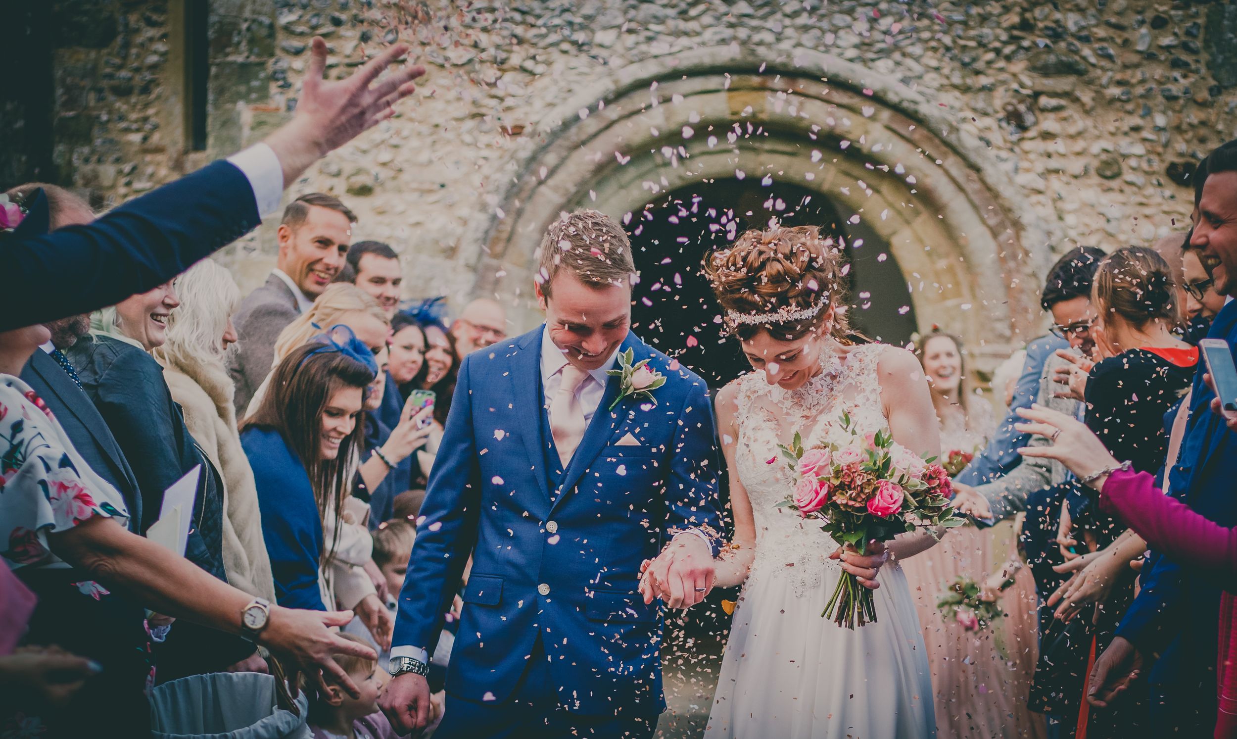 Wedding ceremony guests throwing confetti in front of historic stone archway.