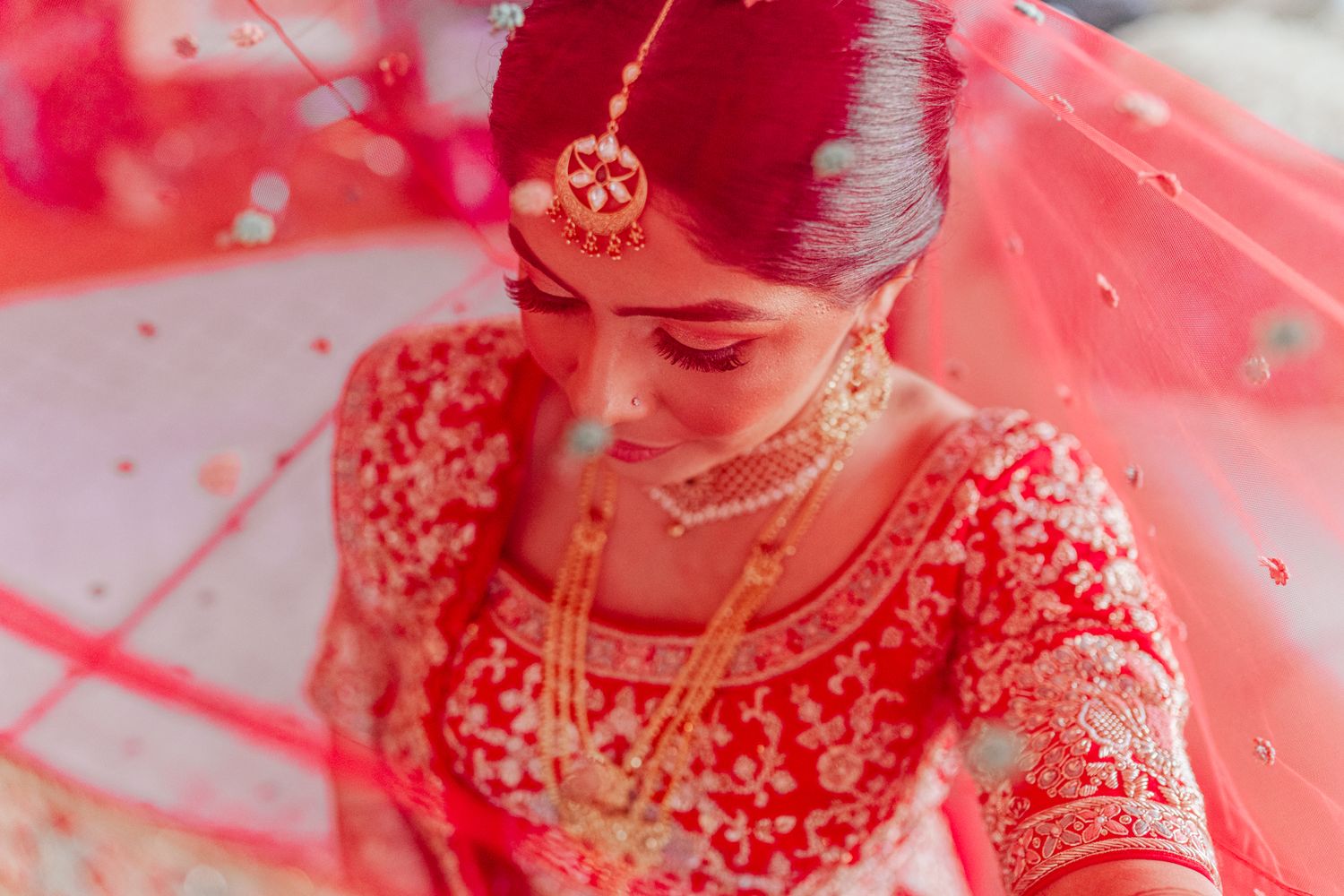 Bride in traditional red lehenga with gold jewelry and embroidery details sits under flowing red veil.
