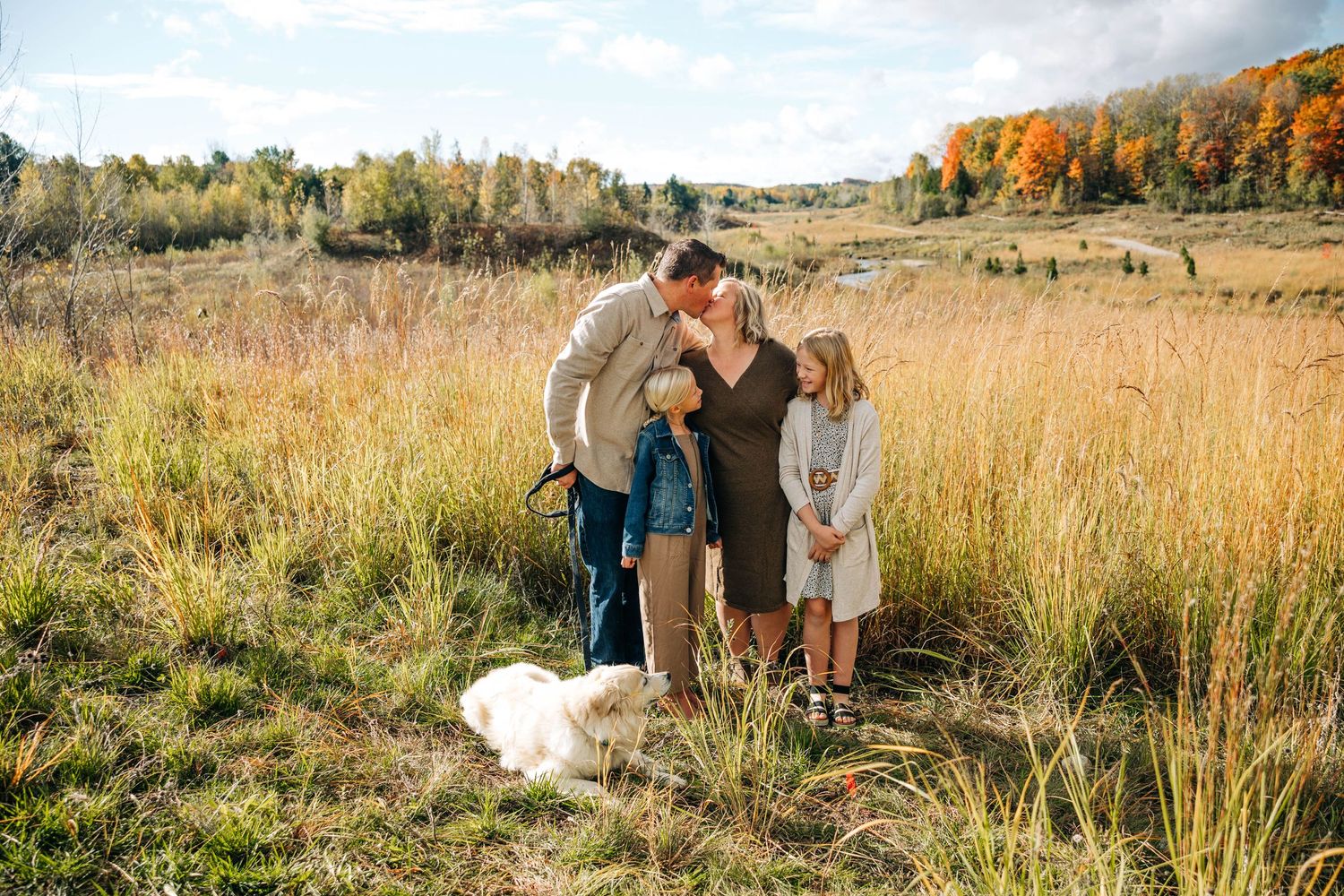 A mom and dad kiss while their daughters laugh with their dog along the Boardman River in the fall
