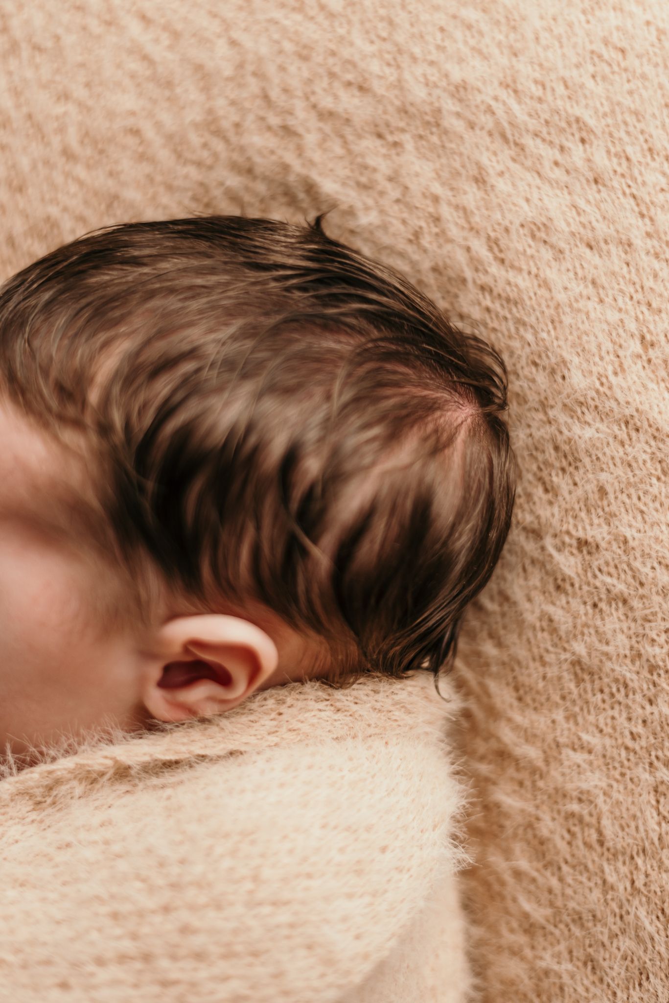 Side view of newborn baby's head with dark hair resting on beige textured blanket.