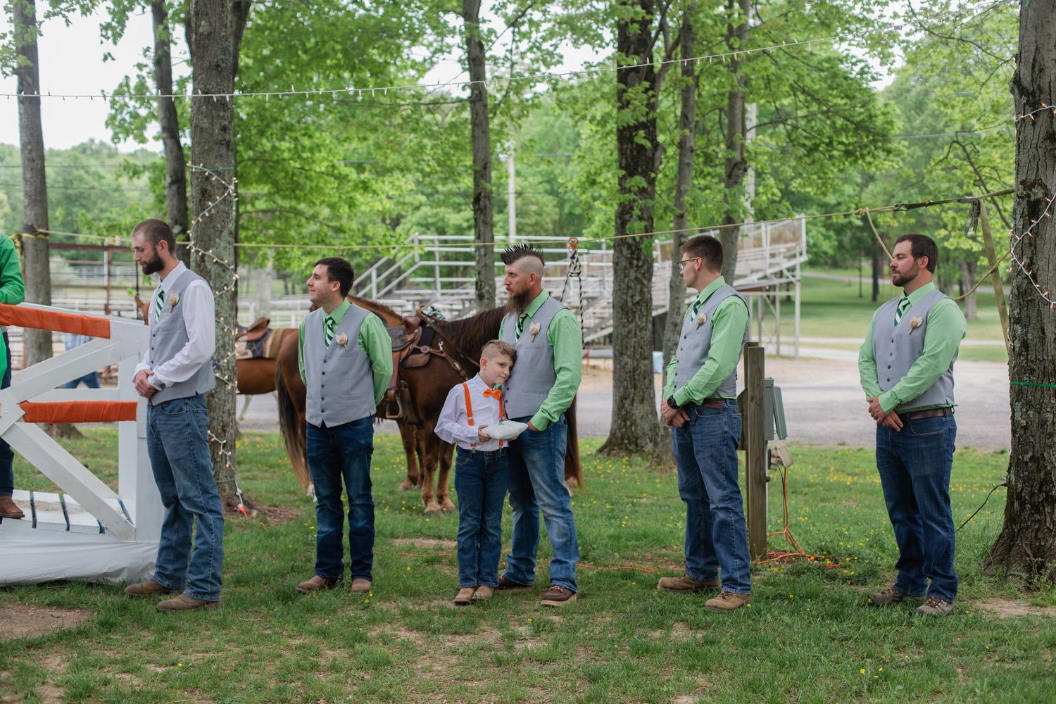 Groomsmen in light gray shirts and jeans stand in a line outdoors during a casual wedding.