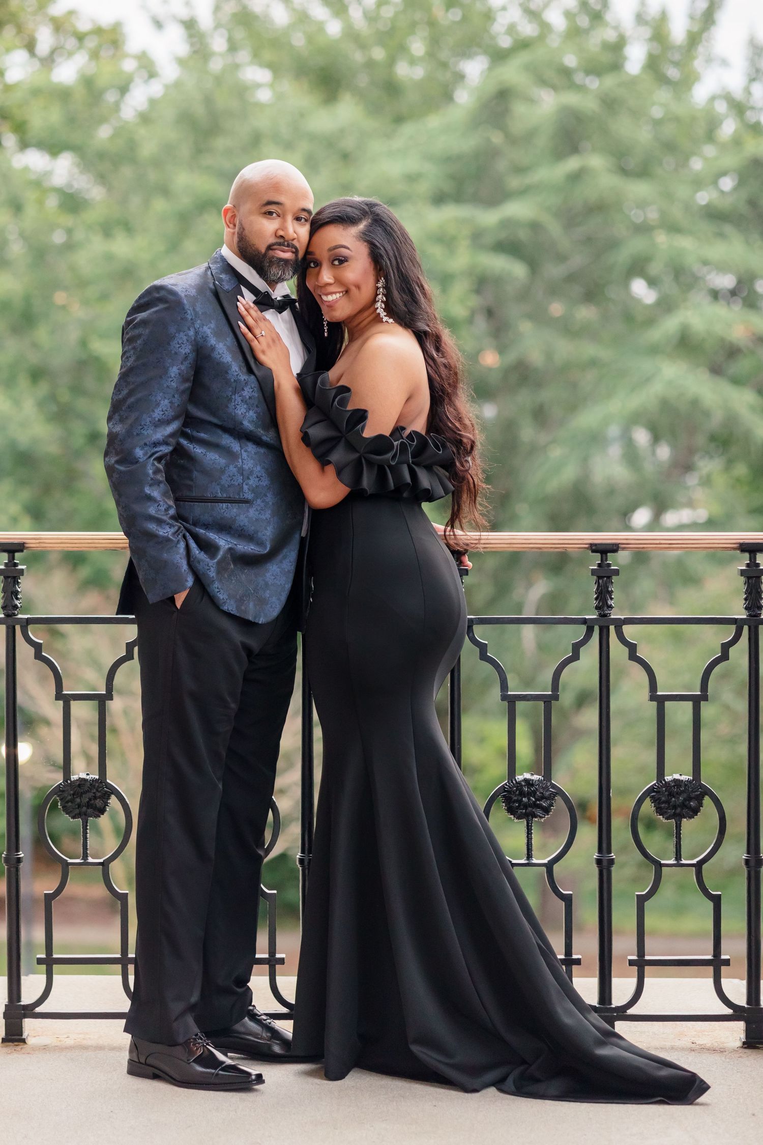 Couple in formal attire pose together on balcony, with woman in black mermaid gown and man in navy suit.