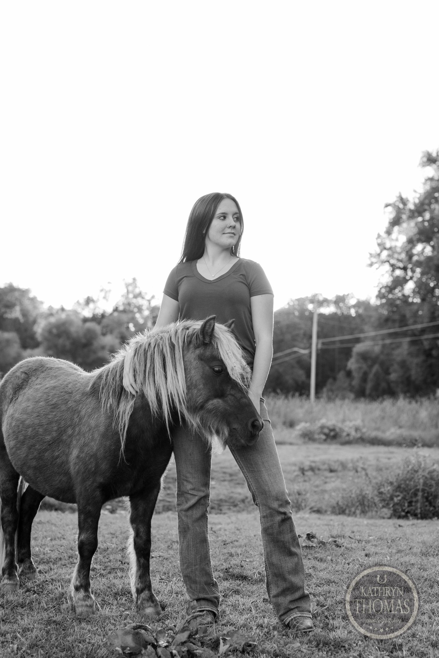 A monochrome portrait of someone standing with a small pony in a rural field setting.