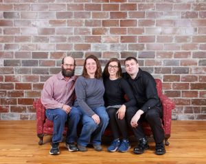 Four adults smiling together on a red sofa against a brick wall with hardwood floors.