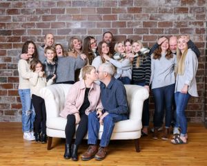 Joyful extended family group photo with elderly couple kissing on white sofa surrounded by loved ones.