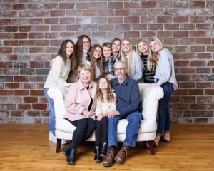 Grandparents seated on white sofa surrounded by granddaughters in a warm family portrait by brick wall.