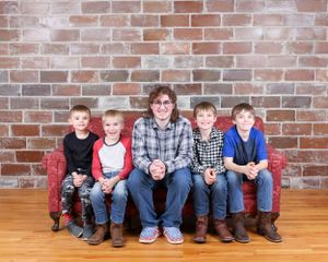Woman sitting on a red sofa with four young boys in a family portrait against a brick wall.