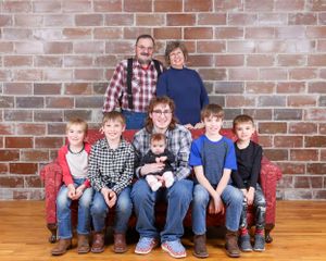 Multi-generational family portrait with grandparents, children, and grandchildren on a red sofa by brick wall.