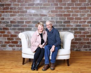 Smiling older couple seated closely together on a white sofa against a rustic brick wall.