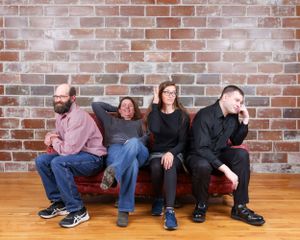 Four adults posing casually on a red sofa against a brick wall in a relaxed family portrait.