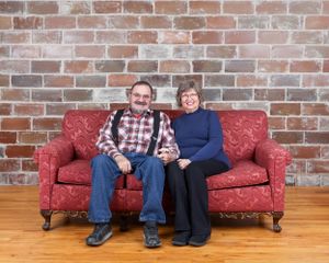 Smiling couple sitting together on a red floral sofa against a rustic brick wall on hardwood floors.