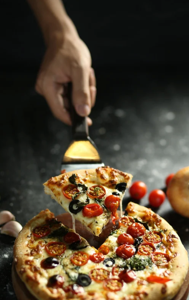 Hand lifting slice of rustic pizza topped with cherry tomatoes, olives and herbs.