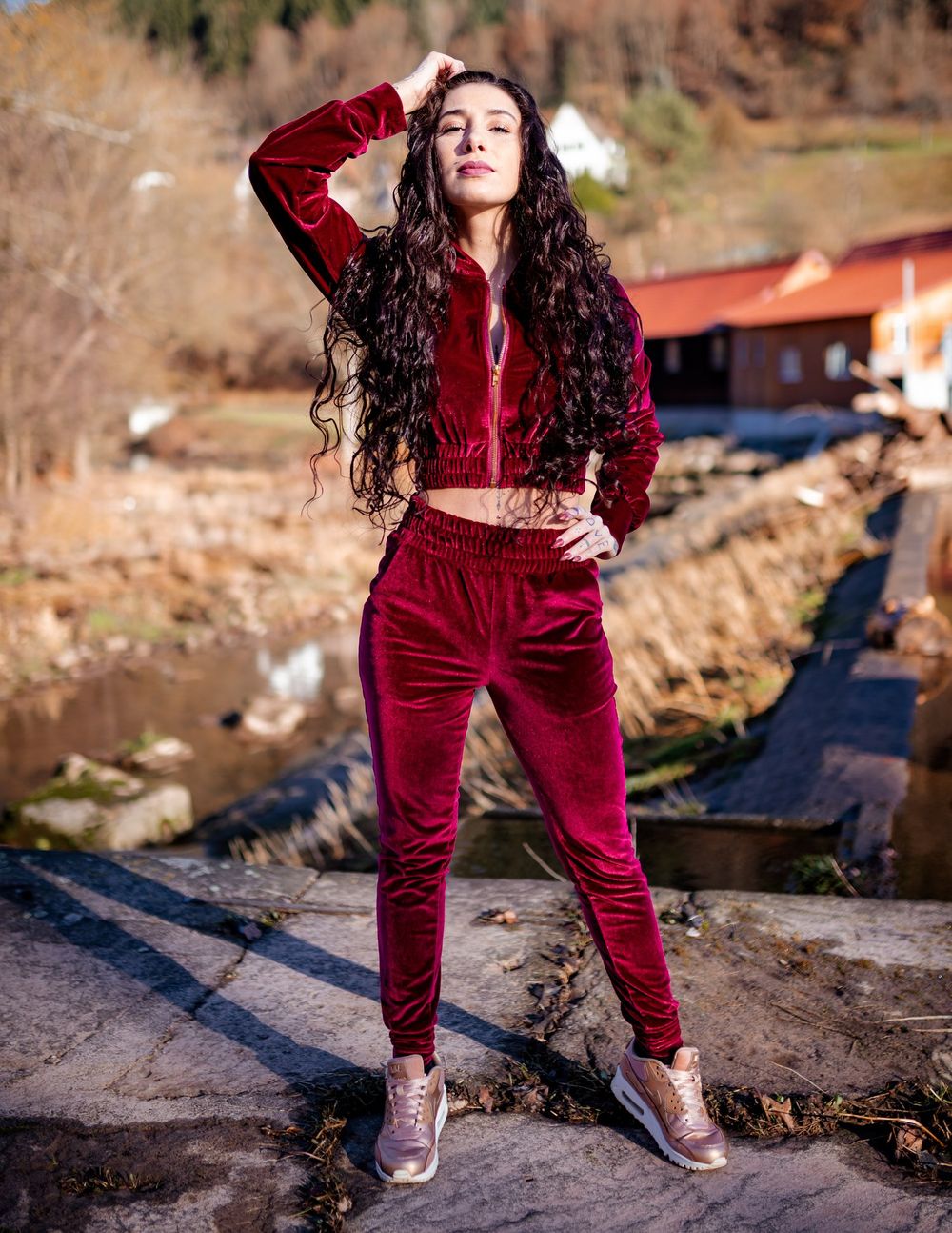 A person in a burgundy velvet tracksuit poses outdoors on a rocky terrain with mountains in the background.