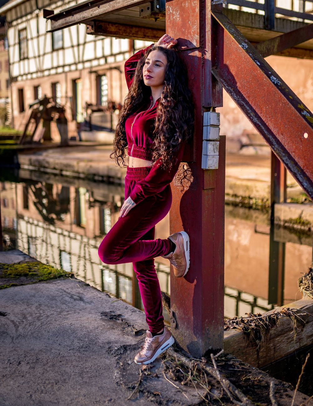 Woman in maroon athleisure outfit poses against metal structure by waterfront at sunset with urban backdrop.