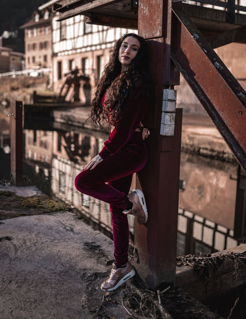 A figure in burgundy velvet lounges against a rustic industrial beam in a moody urban setting.