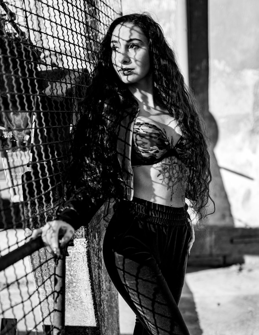 Black and white portrait of model with long dark hair posing against chain link fence in urban setting.