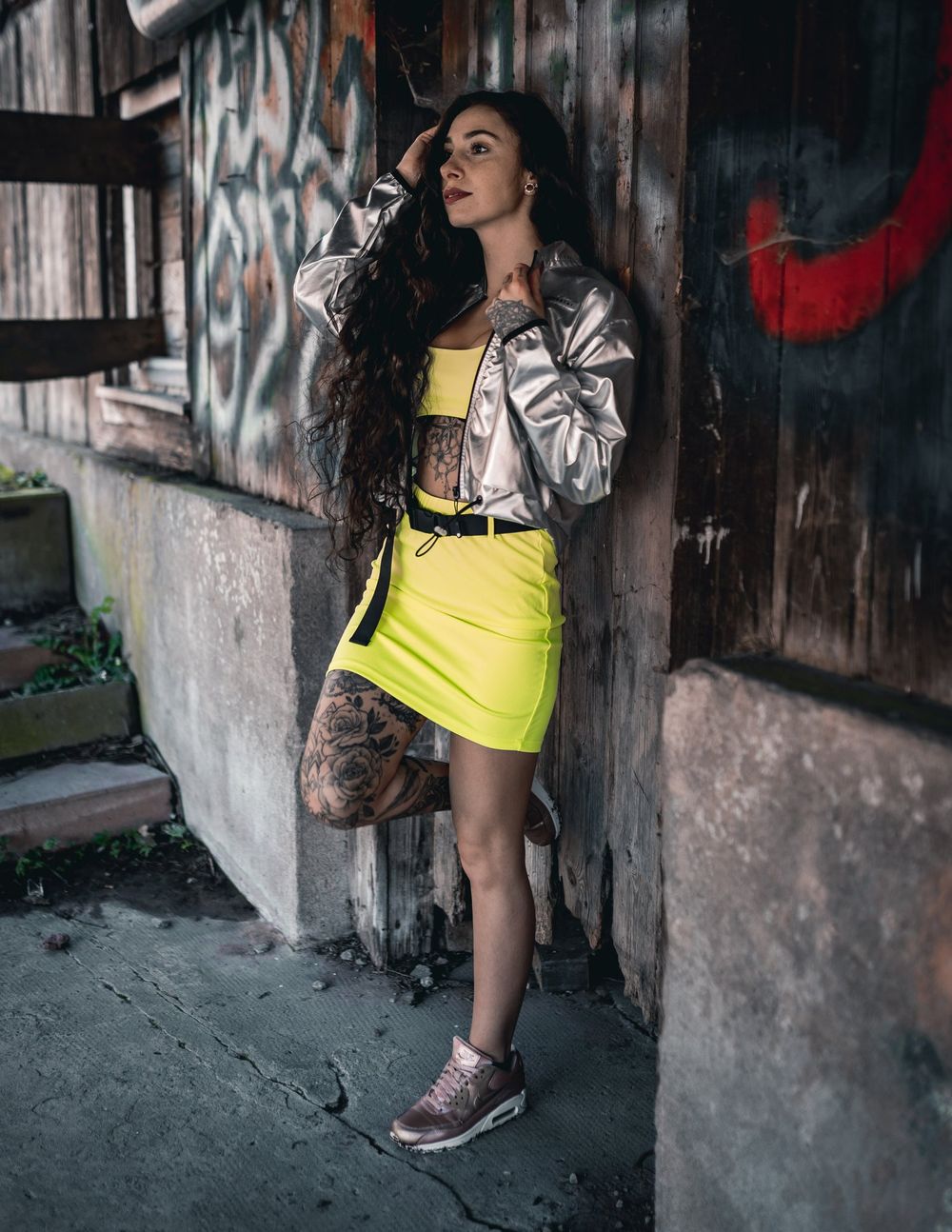 Street style fashion model wearing silver jacket and neon yellow skirt against graffiti wall.