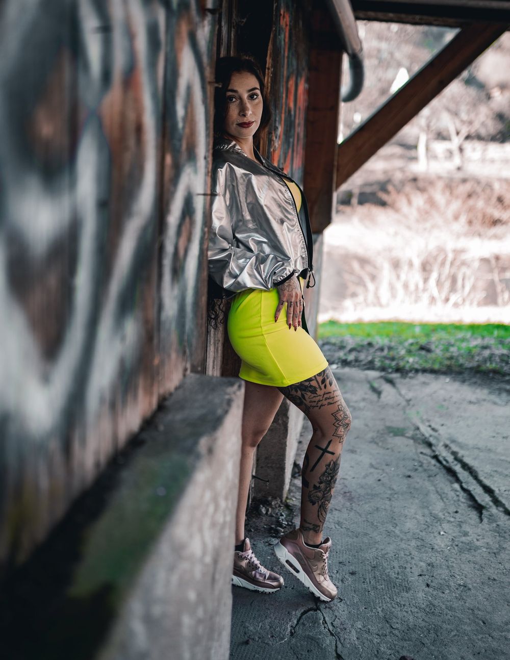 Fashion model in metallic jacket poses against urban graffiti wall wearing neon yellow outfit.
