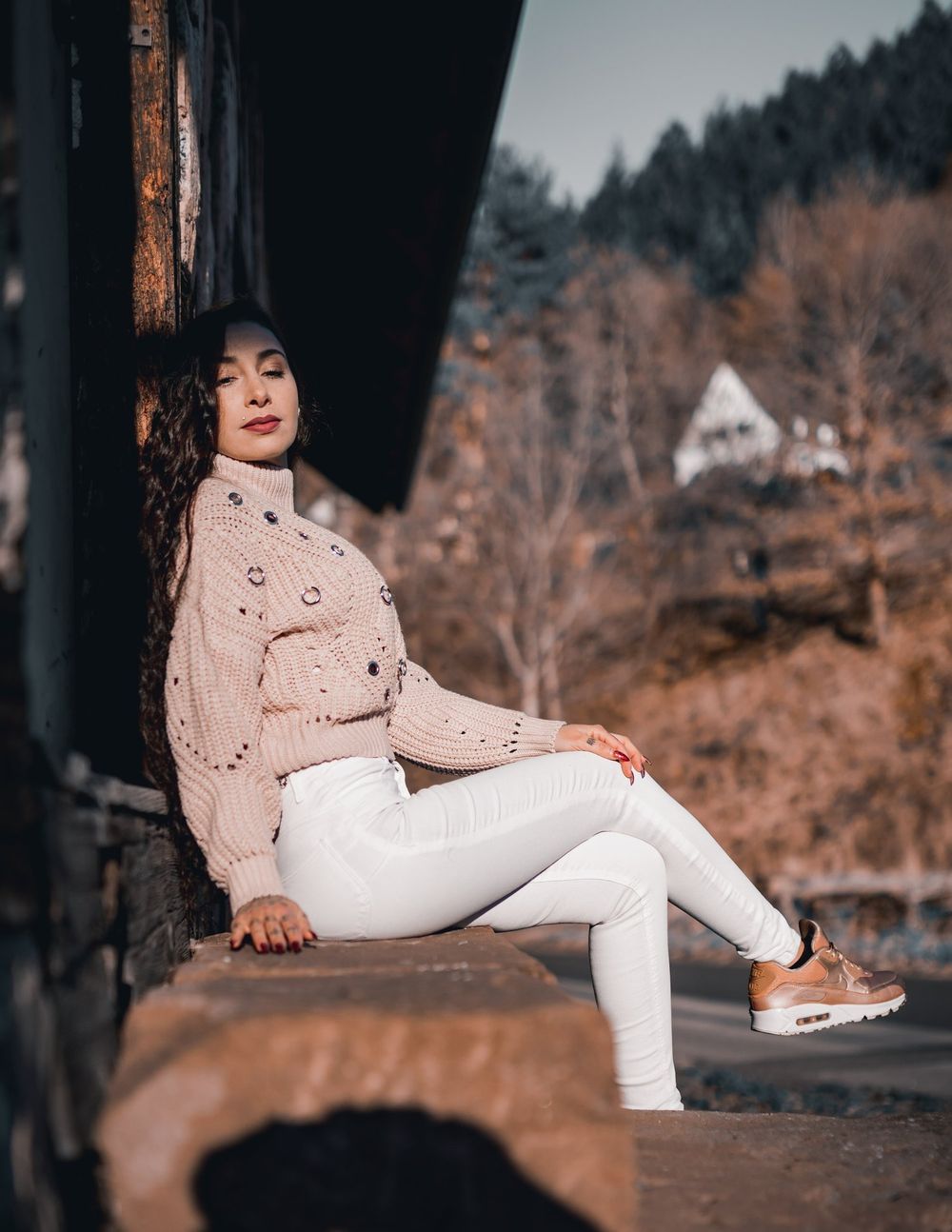 A fashion photoshoot featuring a pink knit sweater and white pants against a rustic outdoor backdrop with autumn trees.