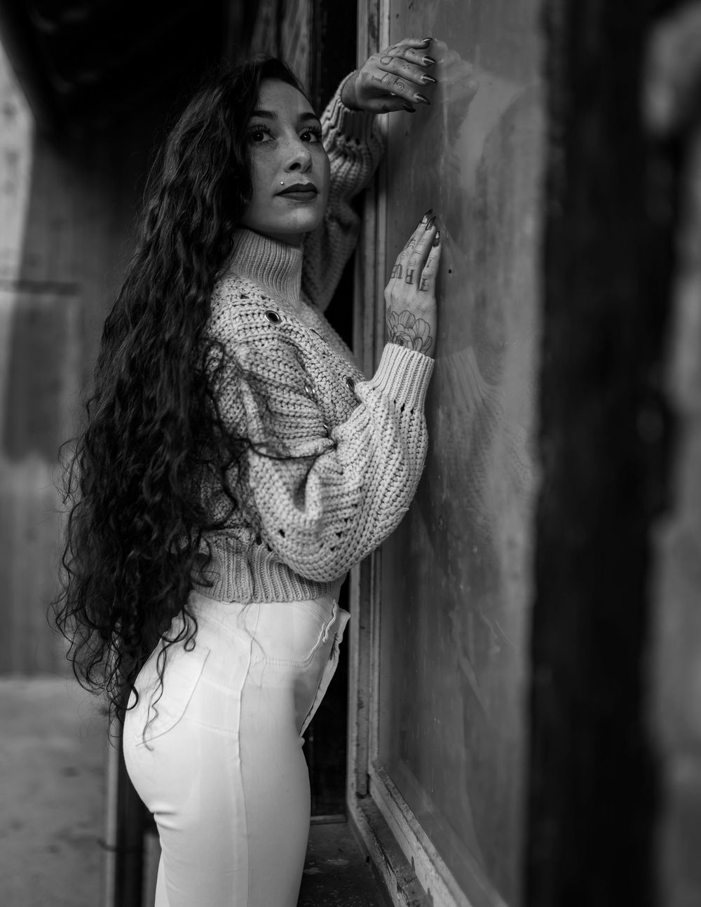 Black and white portrait showcasing long wavy hair and knit sweater against textured wall.