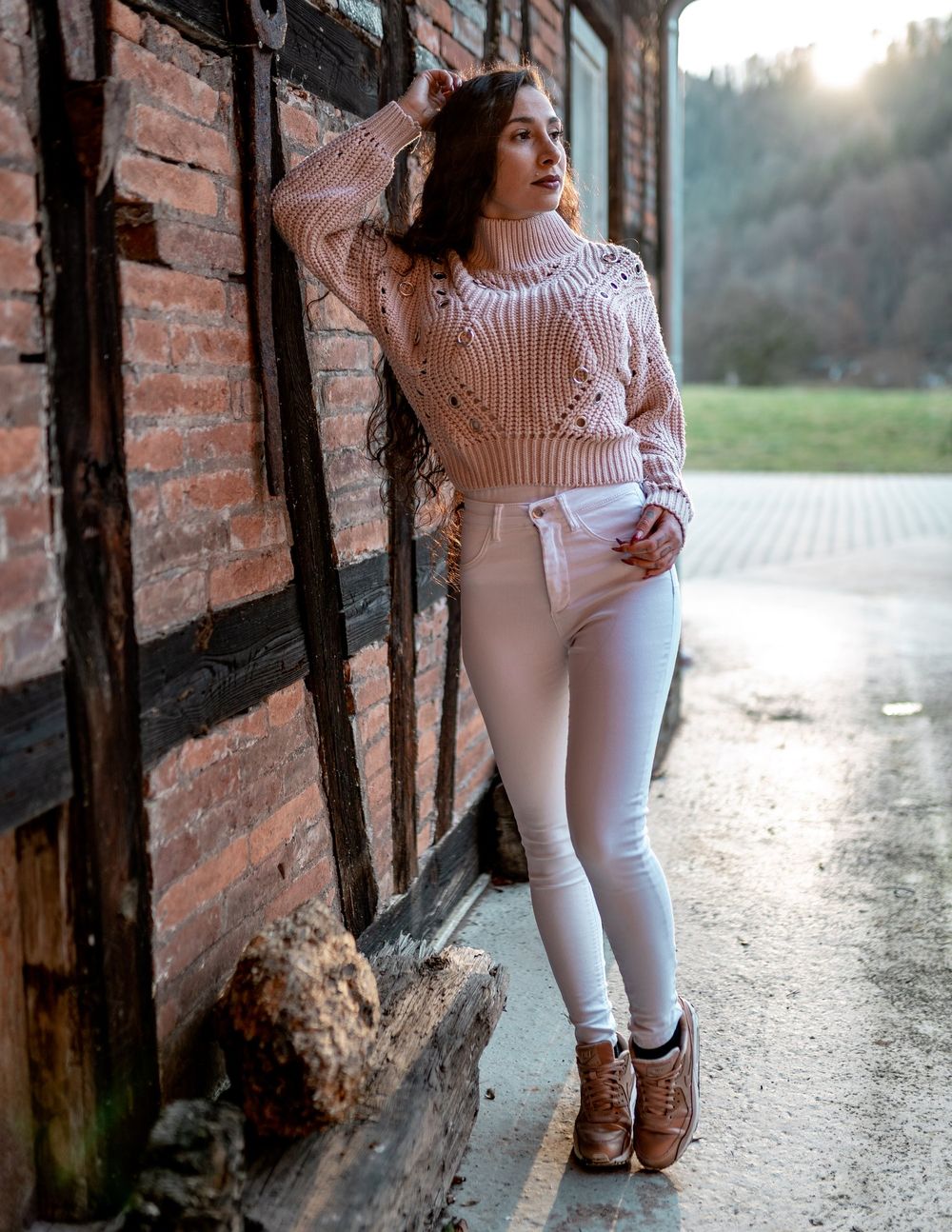 Fashion photography featuring pink cable knit sweater and white jeans against brick wall at sunset.
