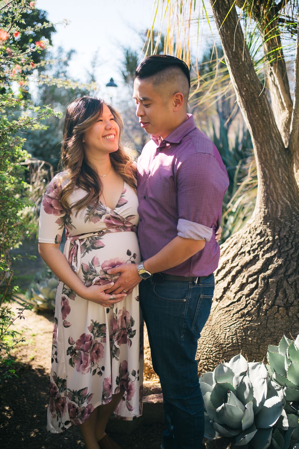Expectant couple in casual attire share an intimate moment under a palm tree in a garden setting.