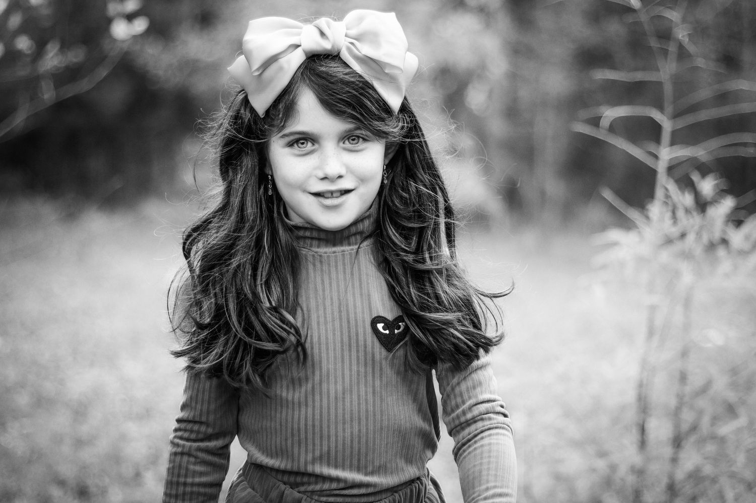 Black and white portrait of a young girl with long wavy hair, wearing a large bow on her head walking toward camera.