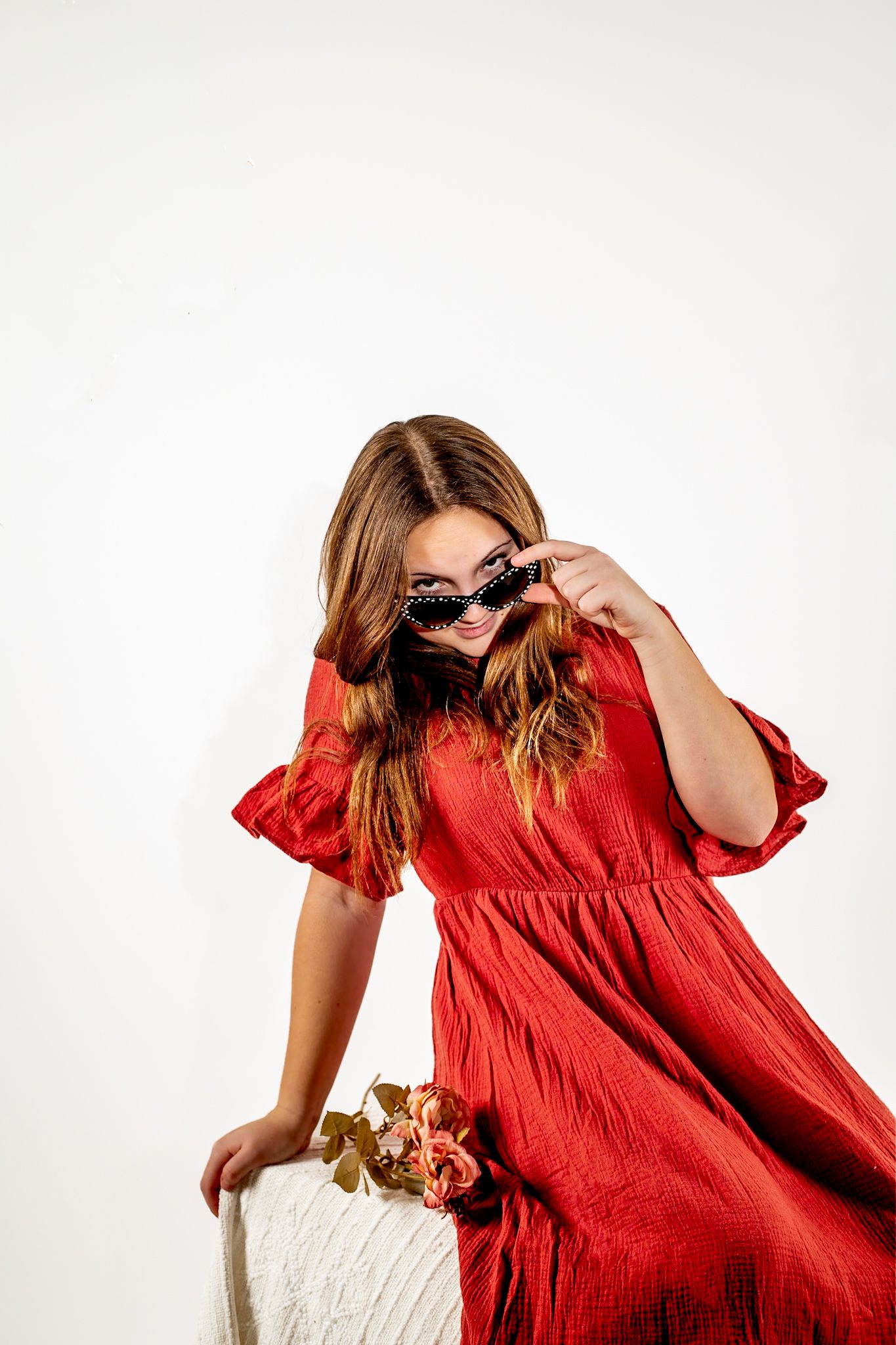 Woman in flowing red dress poses with sunglasses against white background.