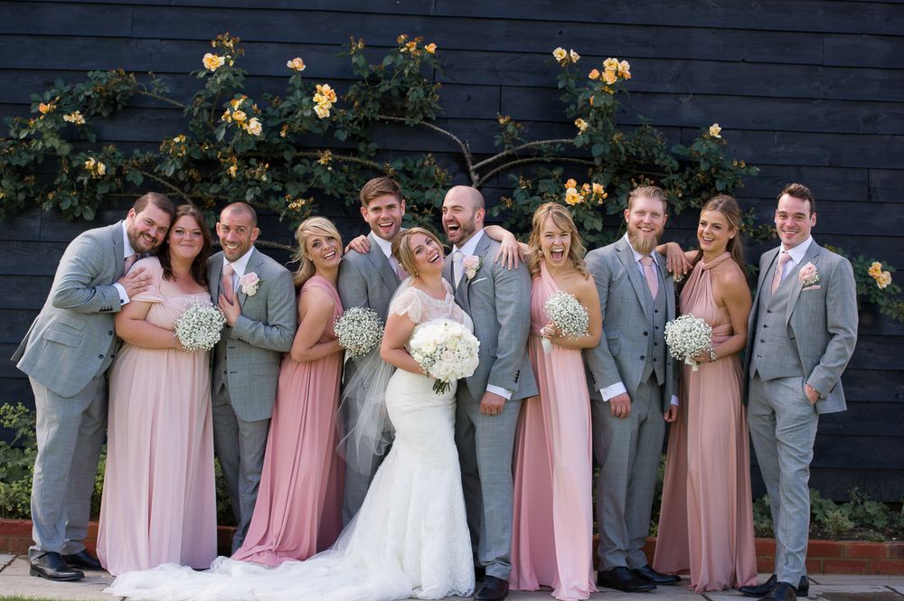 Wedding party in gray suits and pink dresses poses together against dark wall with yellow roses.