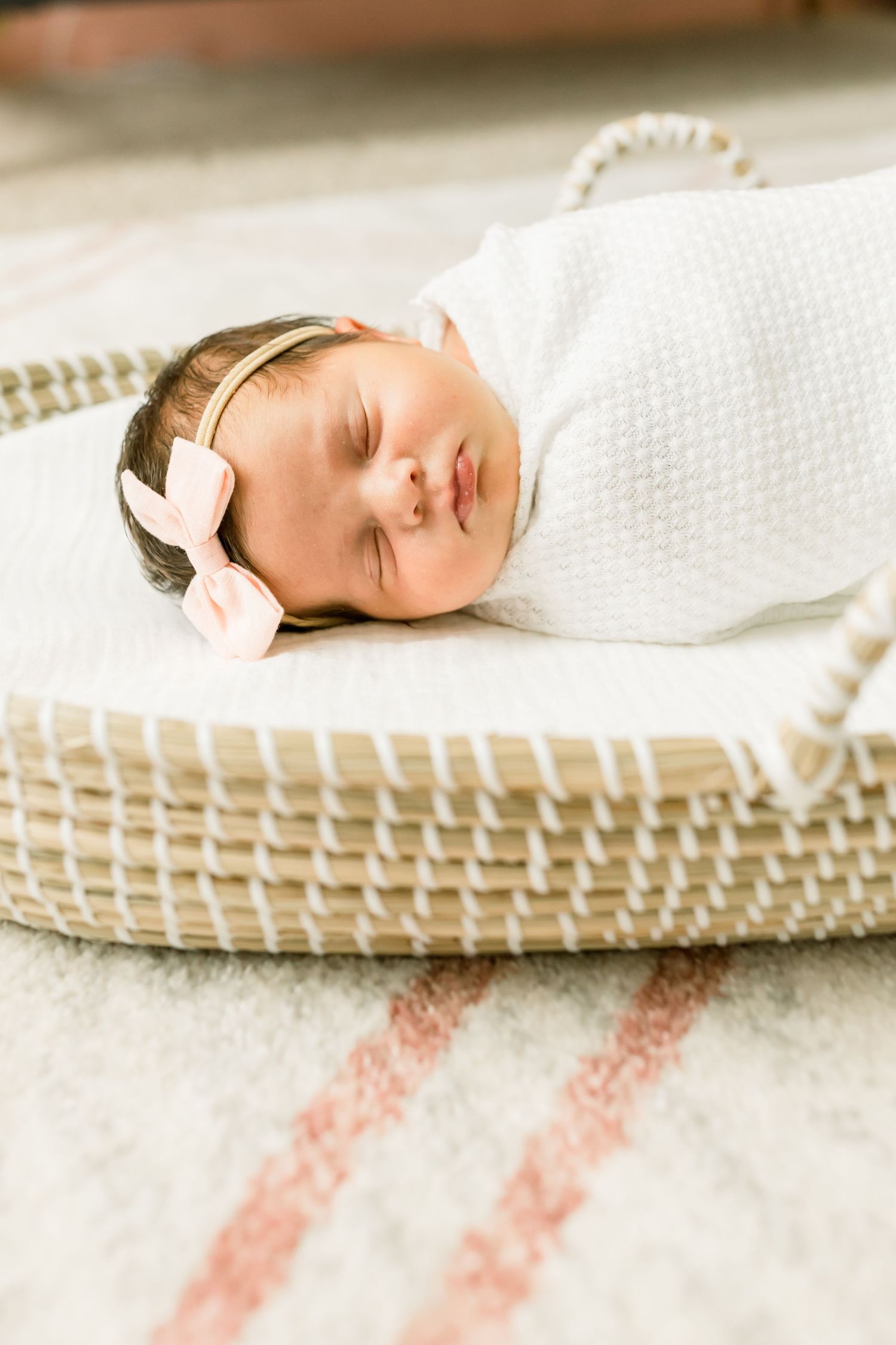 Sleeping newborn wrapped in white swaddle rests peacefully in woven basket on patterned rug.