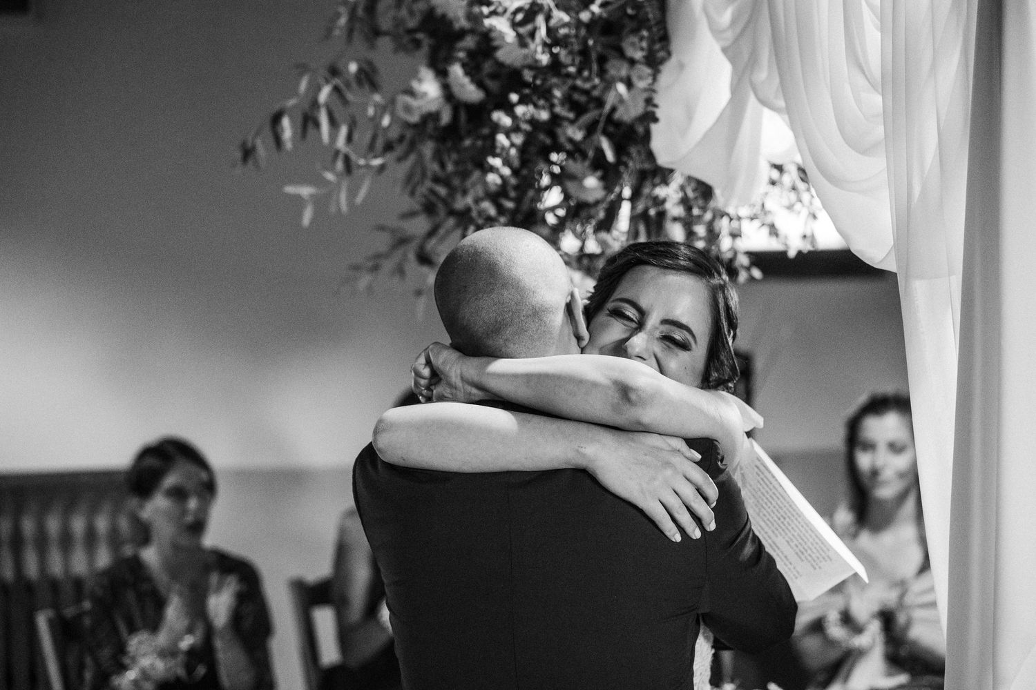 A tender embrace is shared during an intimate black and white wedding moment with onlookers in the background.