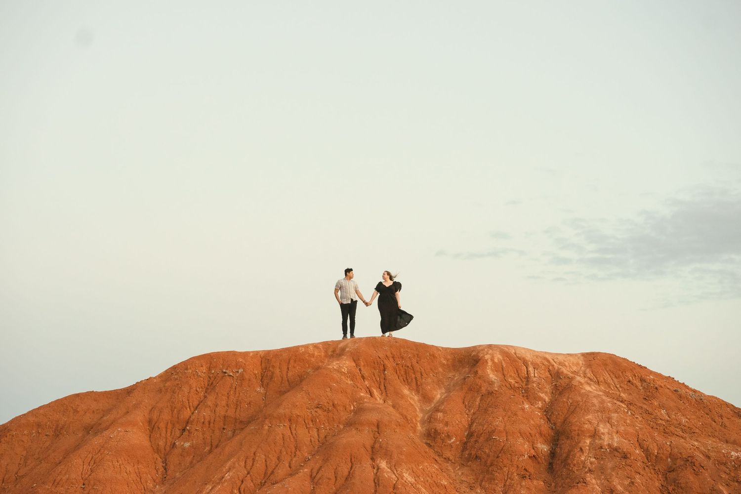 a couple looking towards each other and holding hands on top of a red mountain with a large blue sky behind them