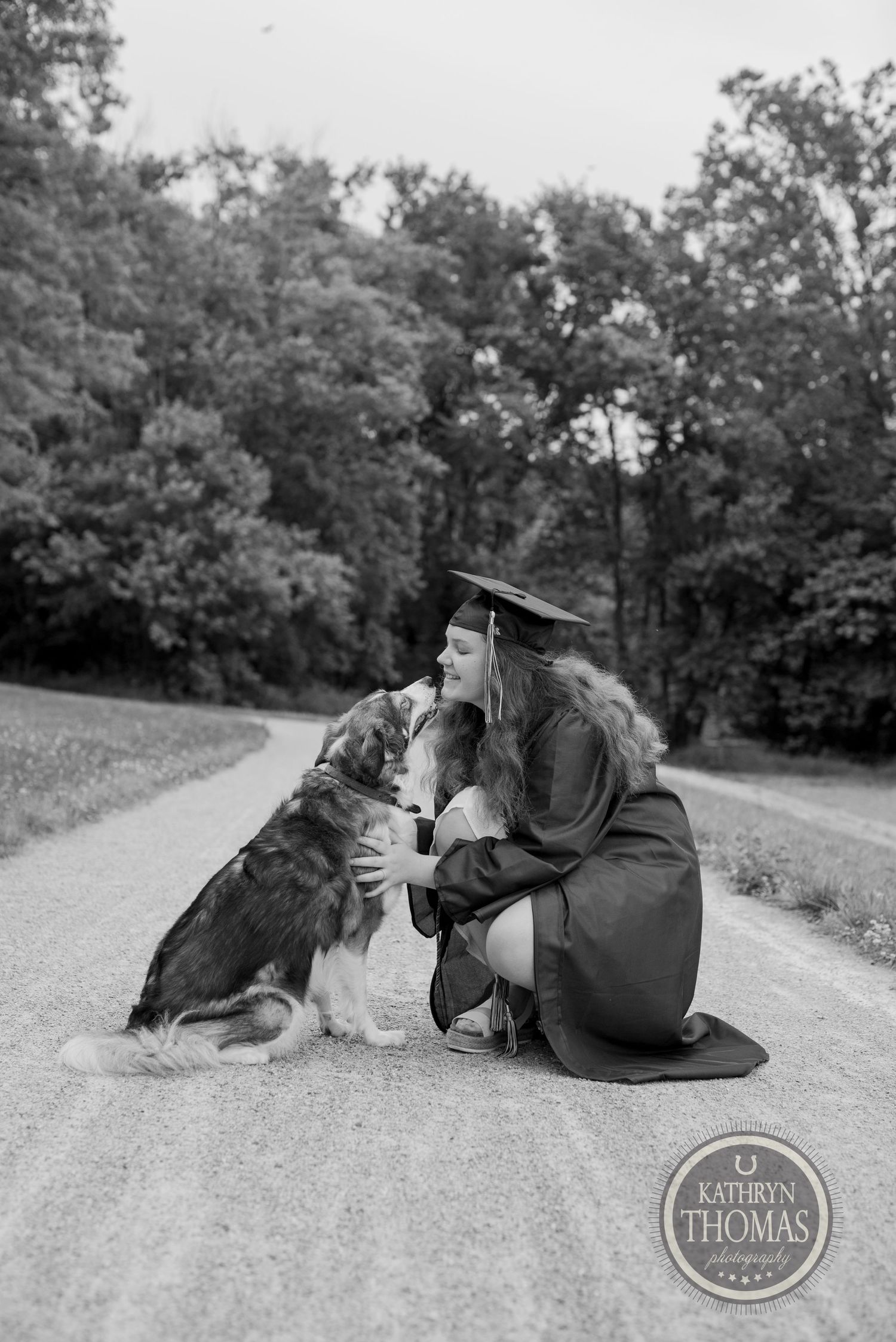 Black and white photo of graduate kneeling on path with dog in academic regalia.