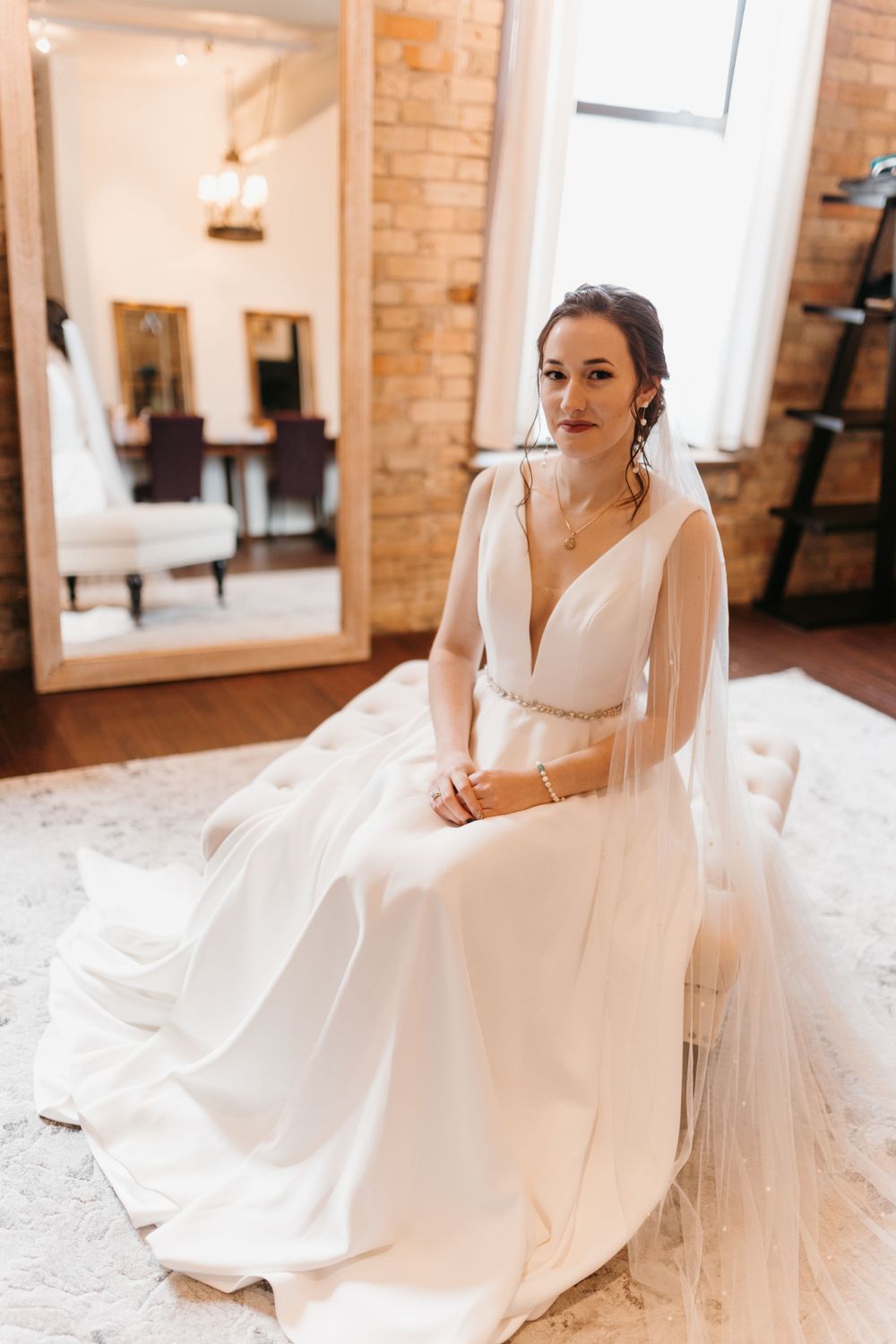 Elegant bride in deep V-neck white gown with beaded belt sits in room with exposed brick walls.