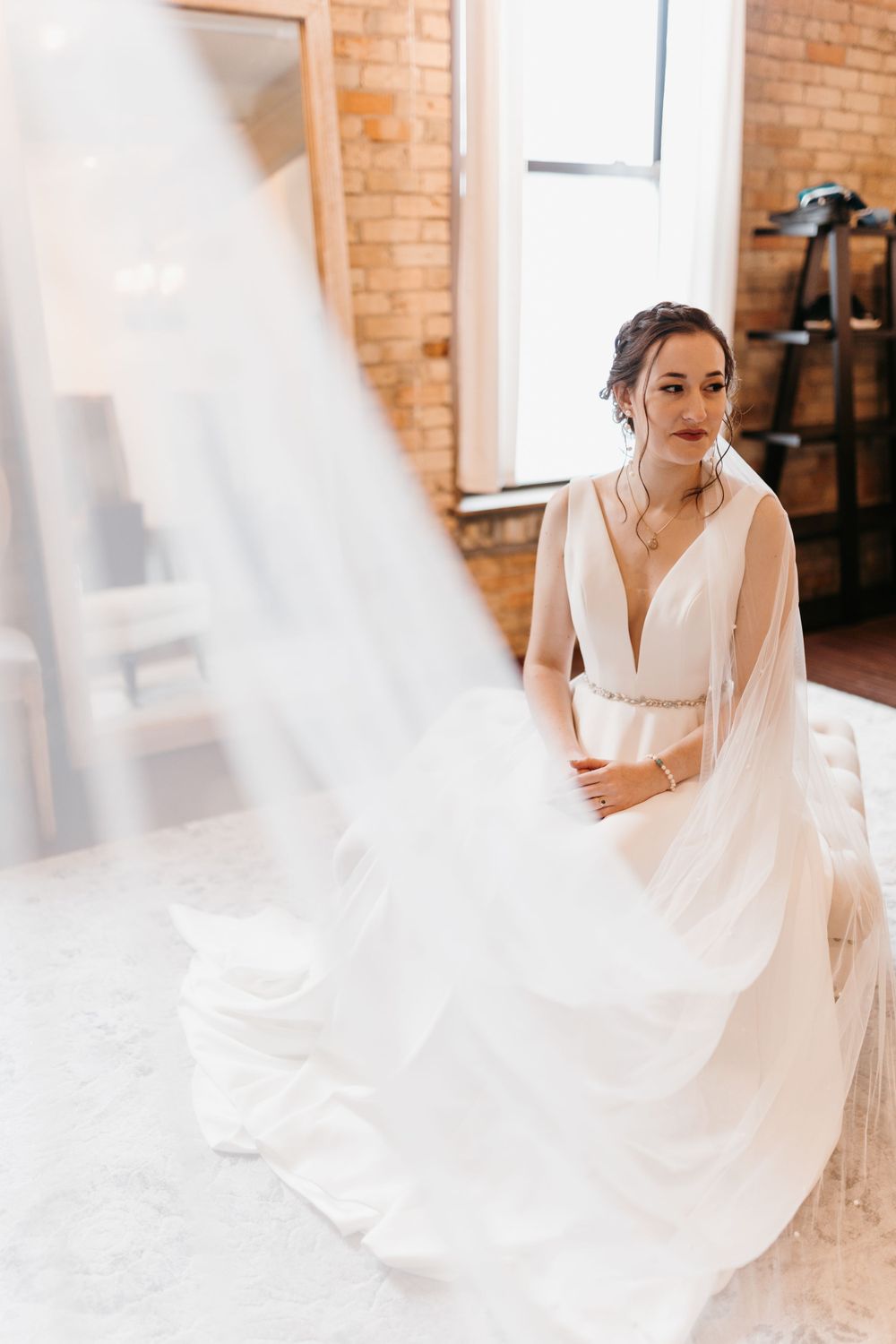 Bride in flowing white wedding gown with veil dramatically sweeping foreground in sunlit brick room.
